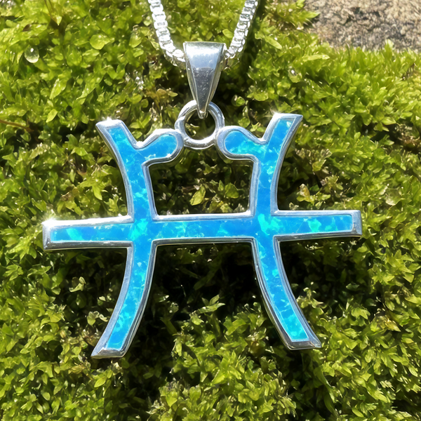 Close-up of the Pisces glyph pendant inlaid with blue opal in a sterling silver setting, on a box chain over moss