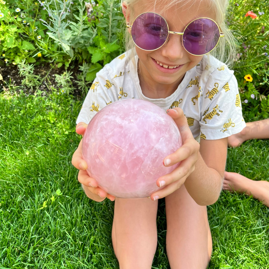A smiling girl in purple sunglasses holding a large rose quartz sphere on the grass