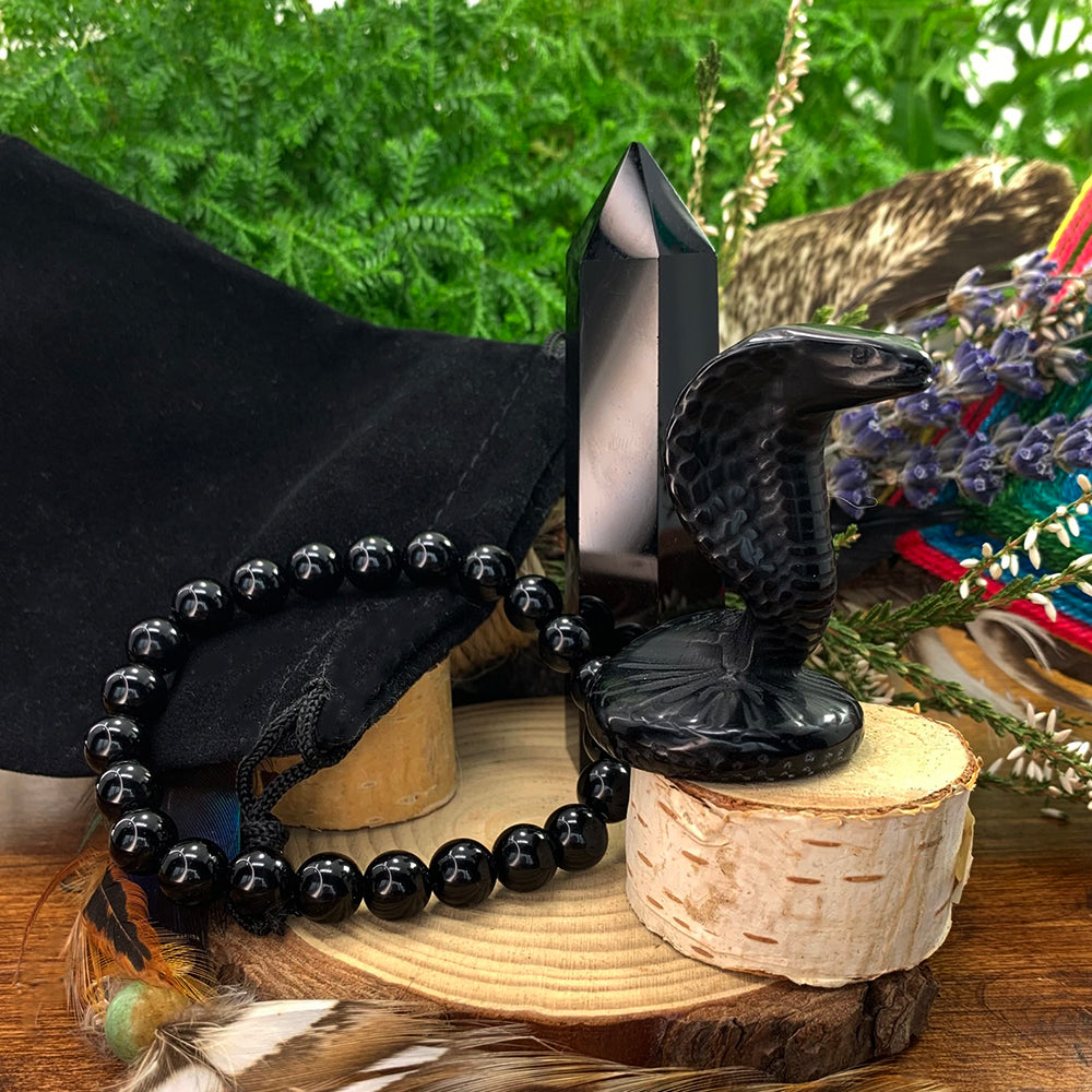 Black agate cobra carving, black obsidian point and obsidian bead bracelet beside a black velvet pouch on birch rounds
