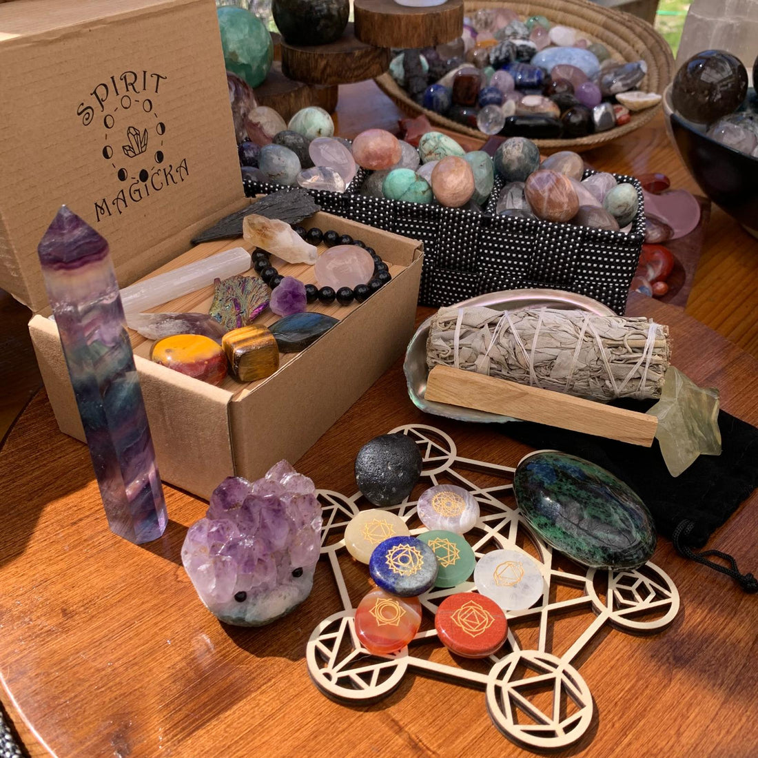 An open Spirit Magicka gift box filled with tumbled stones beside a fluorite point, a sage bundle, a chakra grid and an amethyst cluster
