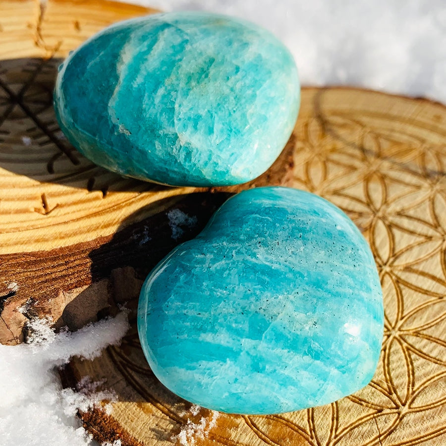 Two polished aqua-blue amazonite hearts with white streaks resting on a carved wooden slab in the snow
