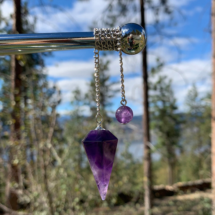 A faceted amethyst pendulum hanging from a silver chain with an amethyst bead, against a forest sky