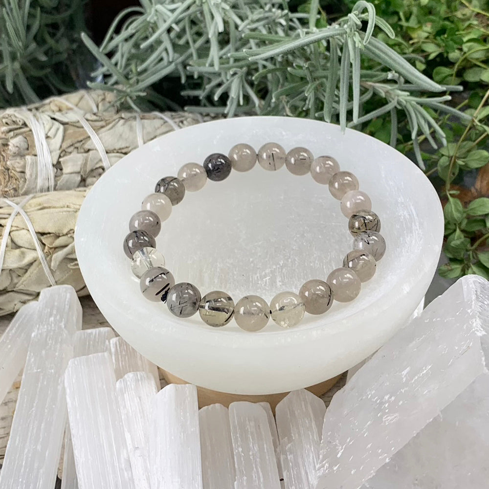A black tourmaline quartz mala bracelet of round smoky-clear beads with black needle inclusions resting in a white selenite bowl