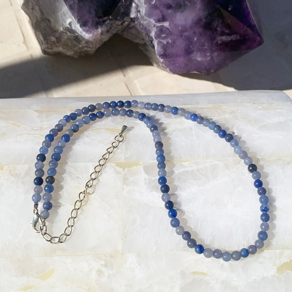A blue aventurine bead necklace in soft and deep blues, laid out with its silver clasp and extender chain on white stone