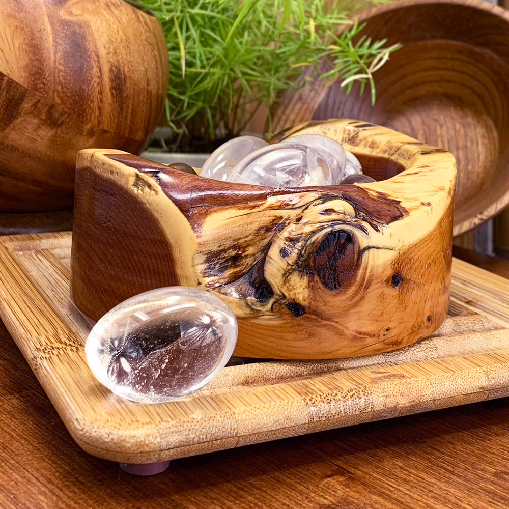 A polished clear quartz ellipse resting on a bamboo tray in front of a carved wooden bowl filled with more clear quartz stones