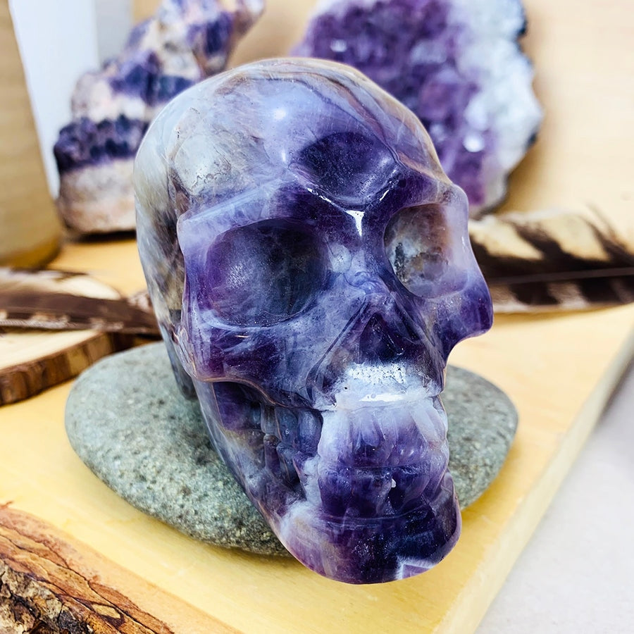 Dream amethyst skull seen face on, resting on a smooth grey stone with amethyst clusters and a feather behind it