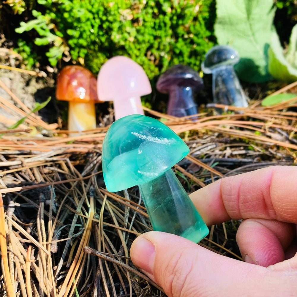 The green fluorite mushroom held between fingers on a bed of pine needles, with other crystal mushrooms behind it
