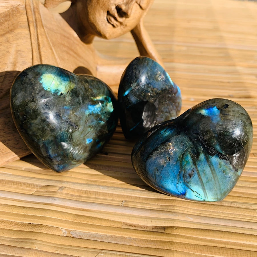 Three labradorite hearts on a woven mat beside a carved wooden figure, each with a different pattern of blue and gold flash