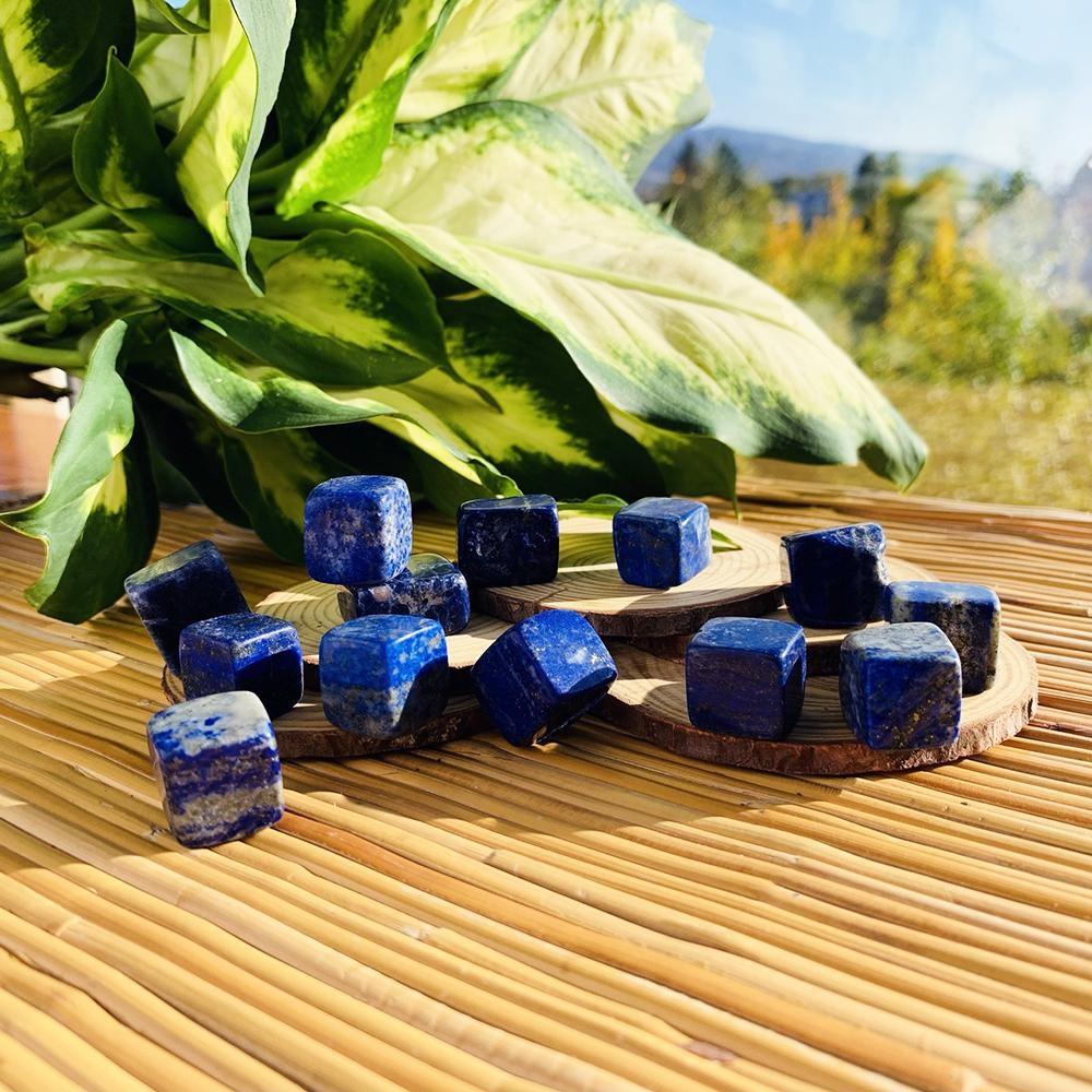 Deep blue lapis lazuli tumbled cubes flecked with gold and white, scattered on wood slices and a bamboo mat beside a leafy plant