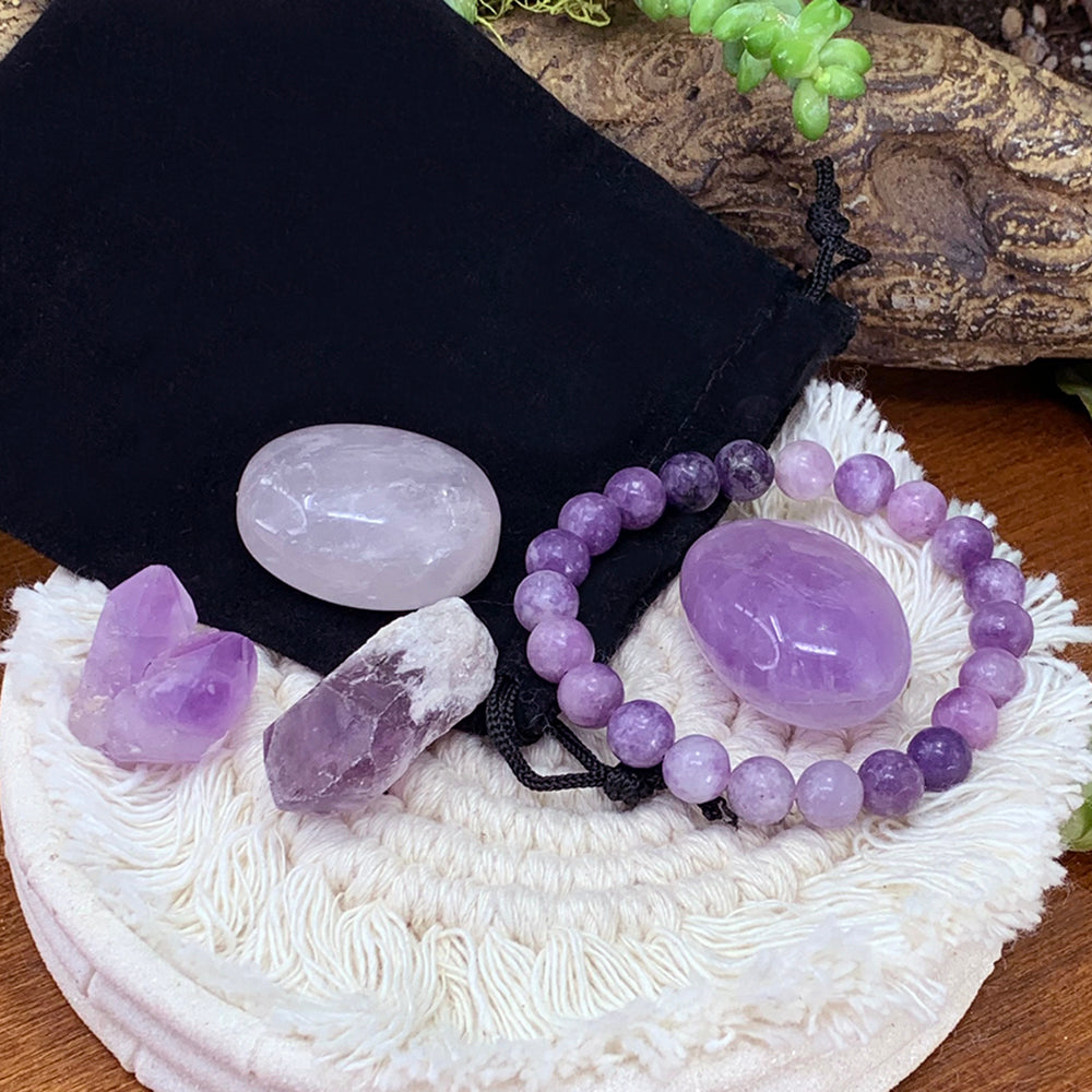 Lavender lepidolite bead bracelet on a black velvet pouch with a purple amethyst tumble, a pale pink rose quartz ellipse, an amethyst shard and a small amethyst cluster