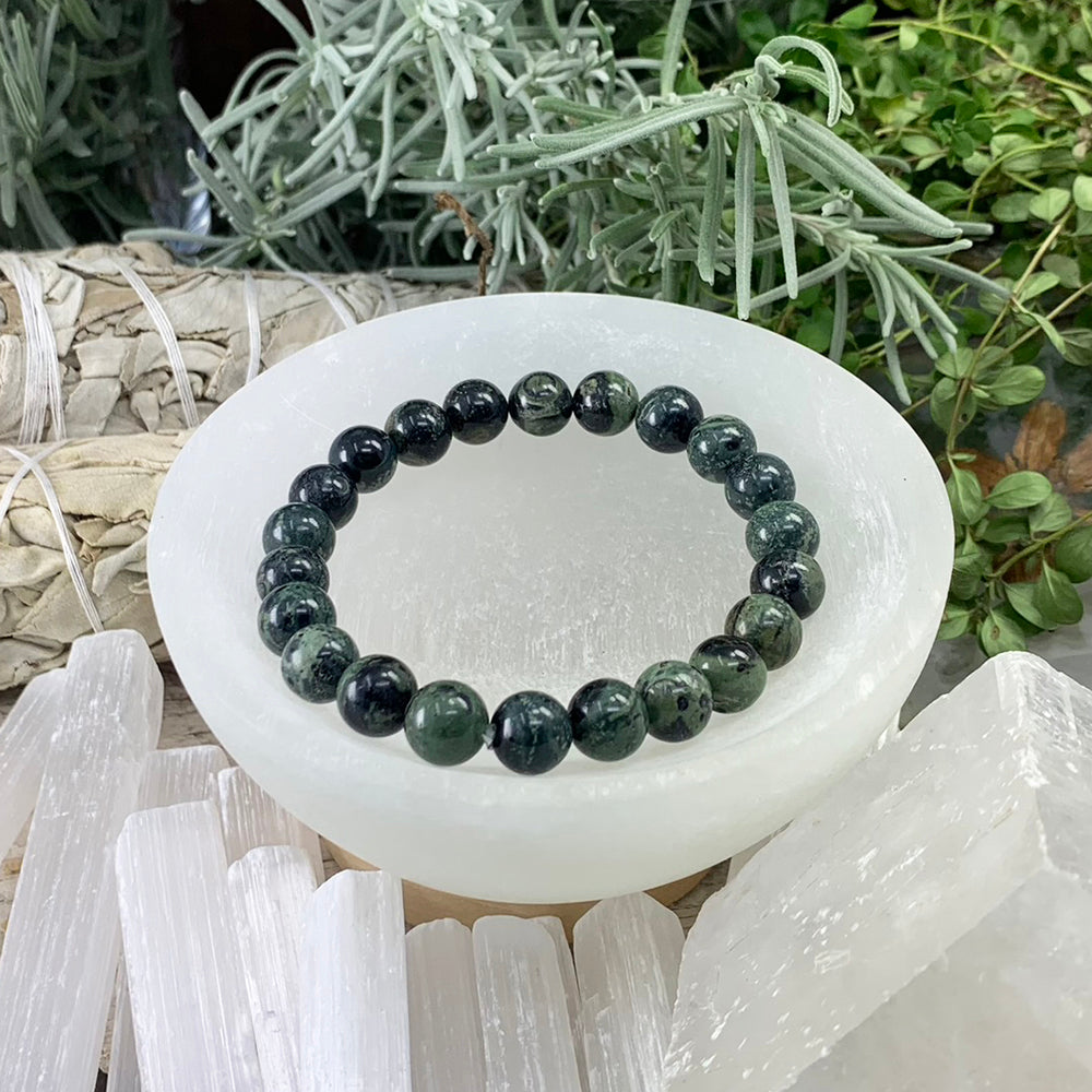 A deep green moss agate bead bracelet resting in a white selenite bowl beside sage bundles