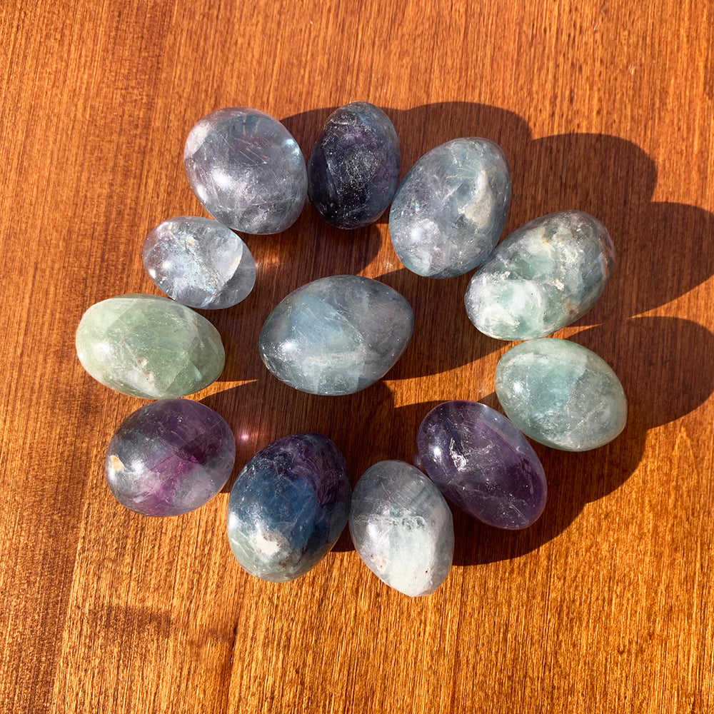 A ring of multicolor fluorite ellipses on a sunlit wooden table, each a different mix of purple, blue, green and clear