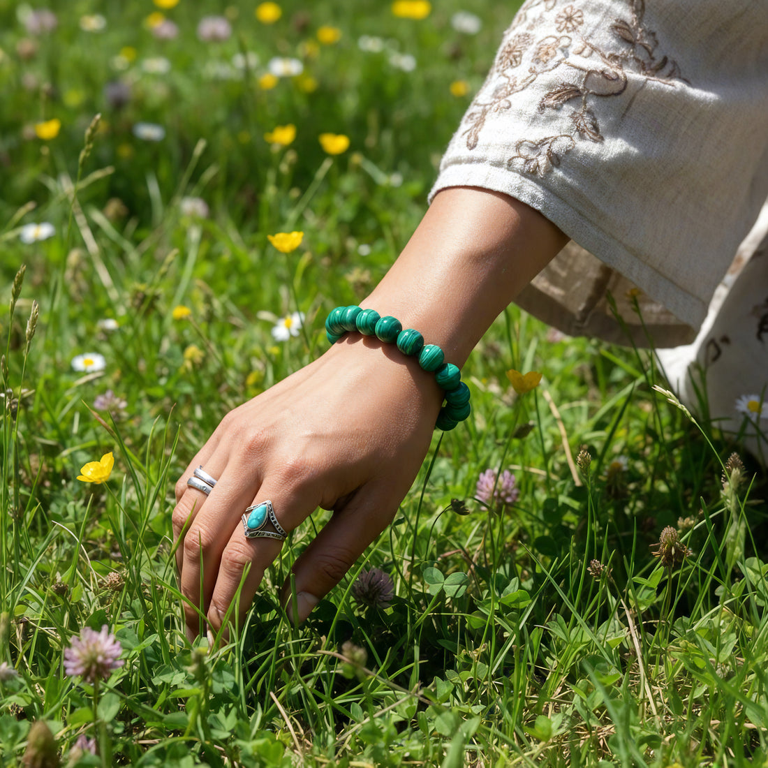 A hand resting in a meadow of wildflowers, wearing the malachite bracelet at the wrist