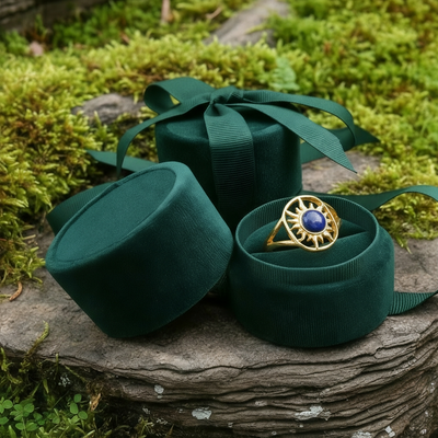 The solar wisdom ring nestled in an open round green velvet ring box beside its lid and ribbon on a mossy stone
