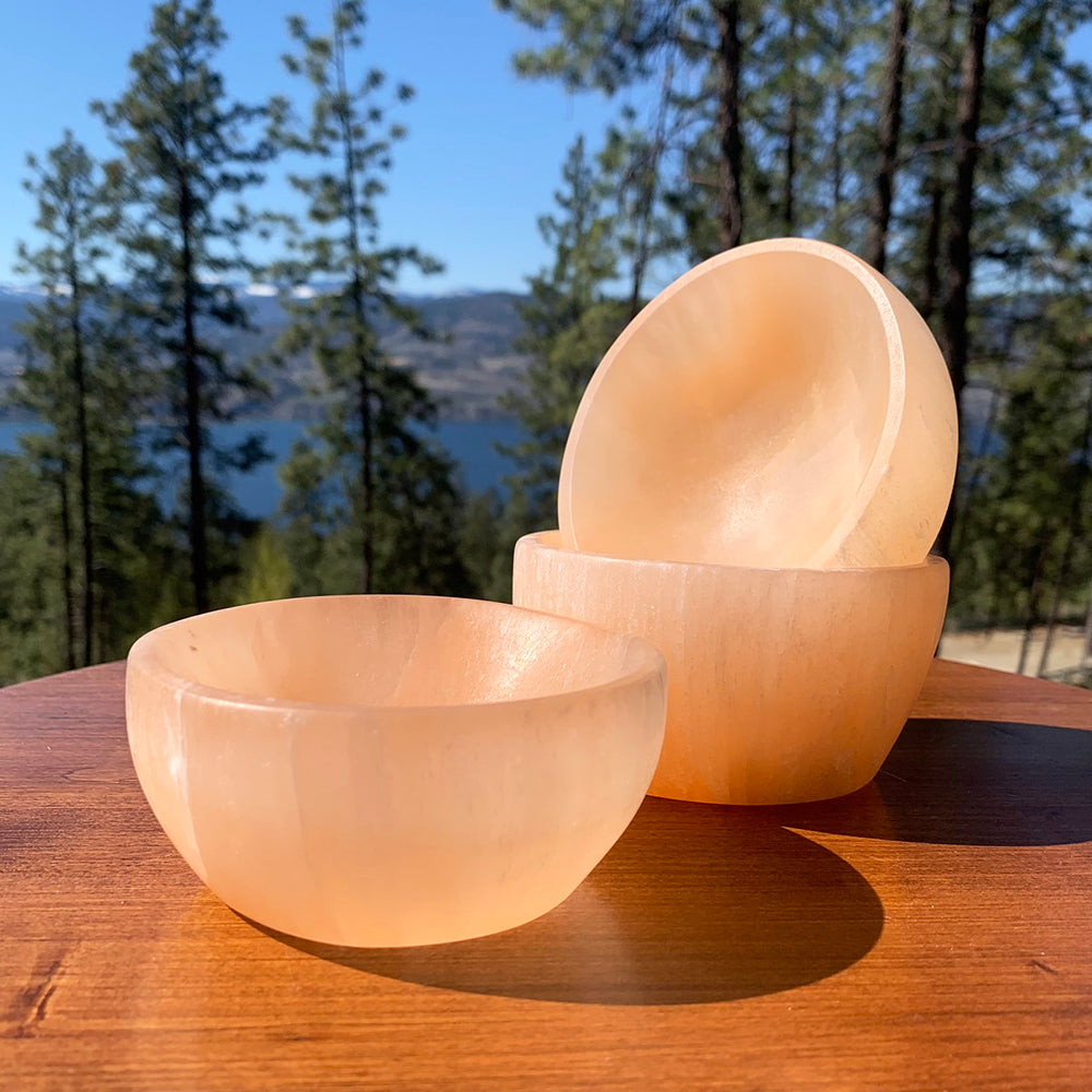Three orange selenite bowls glowing peach in the sun on a wooden table, with pines and a lake behind