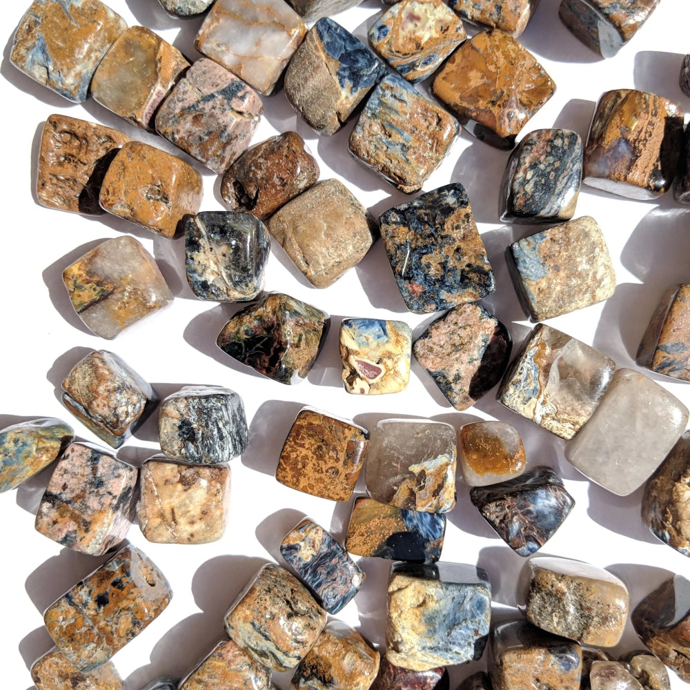 Dozens of pietersite tumbled stones on a white surface in mixed gold, copper, brown, grey and blue tones