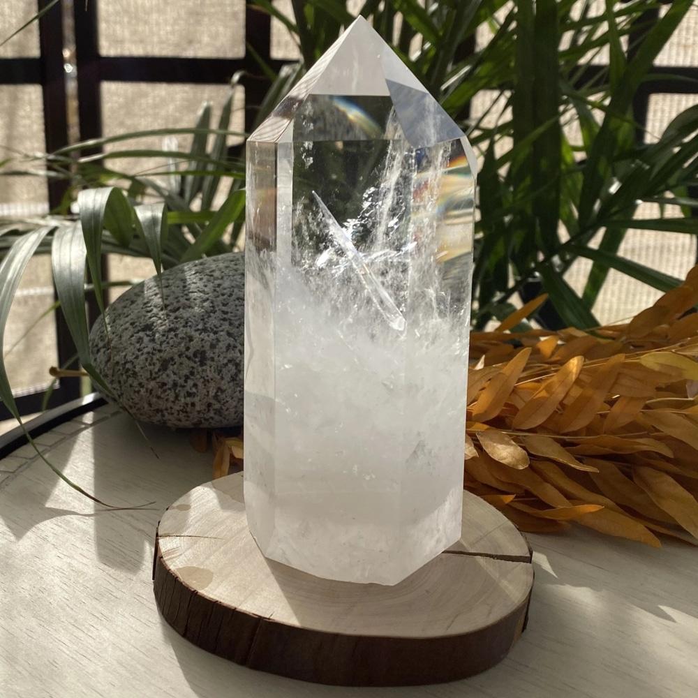 Clear quartz generator standing on a wooden slice in sunlight, its clear faceted tip fading into frosted white veils at the base