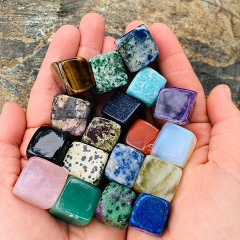 Two cupped hands holding nineteen colourful tumbled crystal cubes, including a red jasper cube