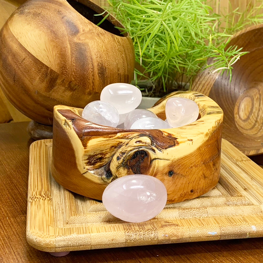 A polished pale pink rose quartz ellipse resting on a bamboo tray in front of a carved wooden bowl of more tumbled stones