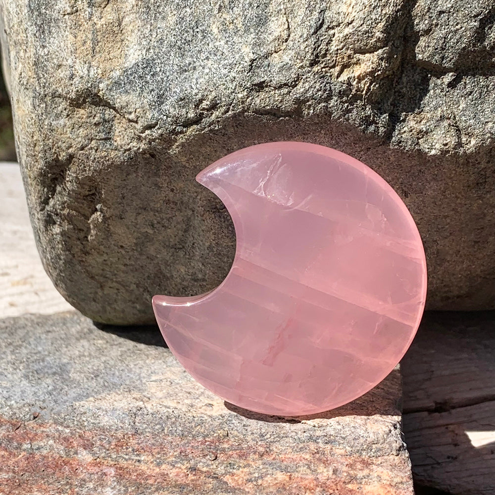 A polished pink rose quartz crescent moon standing upright against a granite boulder