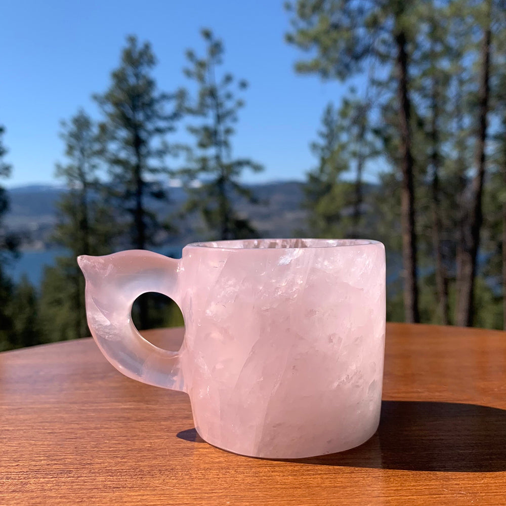 A pale pink rose quartz demitasse cup with a carved leaf-shaped handle on a wooden table, with pines and a lake behind