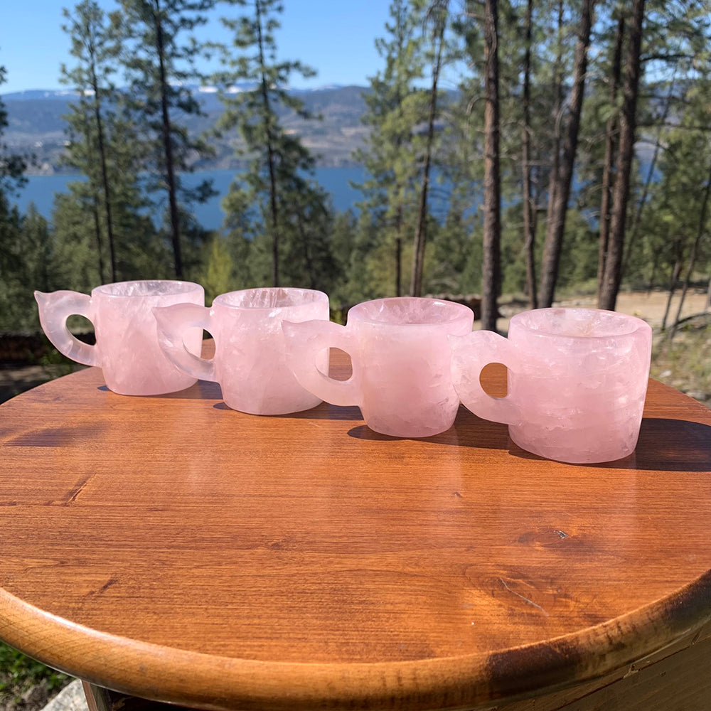 A row of four rose quartz cups on a wooden table, each a slightly different shade of pink and shape of handle