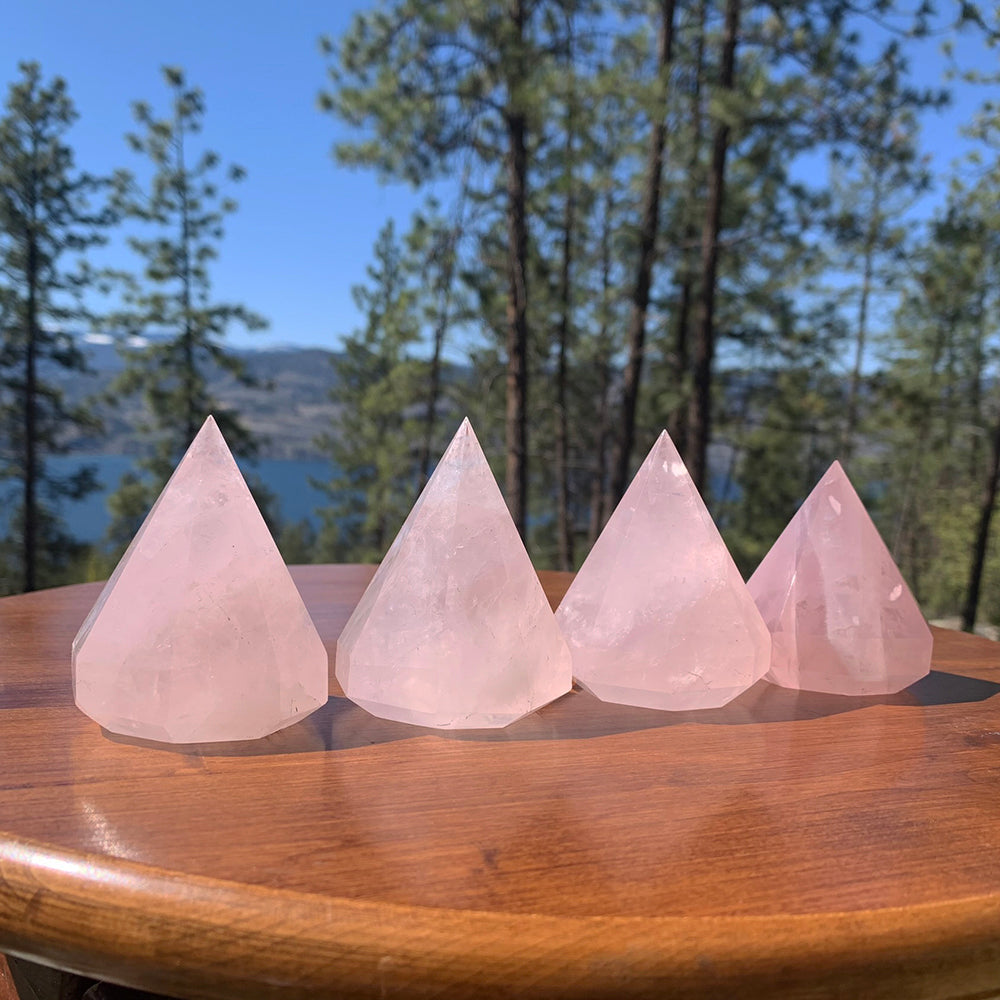 Four faceted rose quartz diamonds standing in a row on a wooden table in the sun, pine trees and a lake behind them
