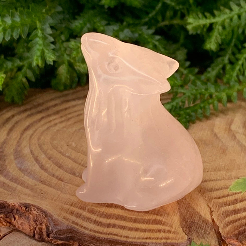 A small carved rose quartz wolf with its head raised, sitting on a wooden slice in front of ferns