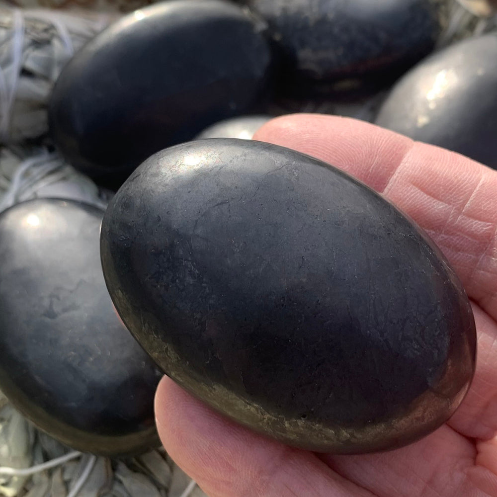 A shungite palmstone resting in an open hand, with more palmstones on bundled sage behind it