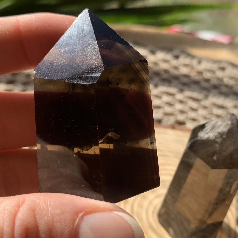 A deep brown, nearly black smoky quartz generator held between finger and thumb, with a lighter one behind it