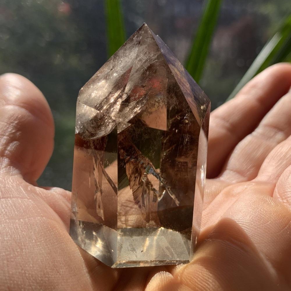 A translucent smoky quartz generator with a pointed tip standing on an open palm in sunlight