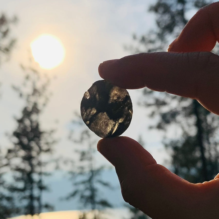 A tektite held between finger and thumb against the sun, glowing smoky brown through its thinner areas
