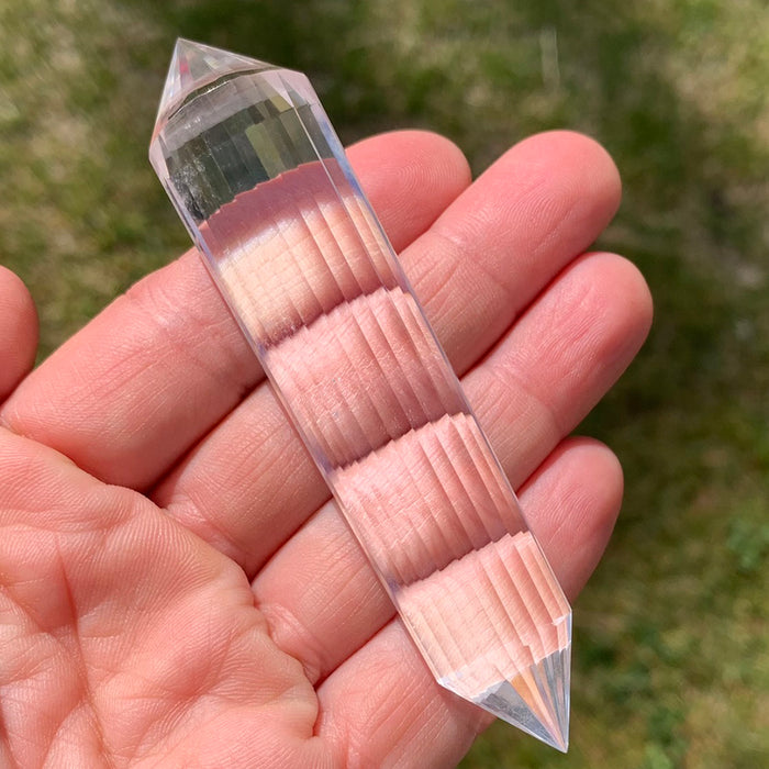 A longer 24-sided Vogel quartz wand lying across an open palm, its narrow facets stacking the light in bands
