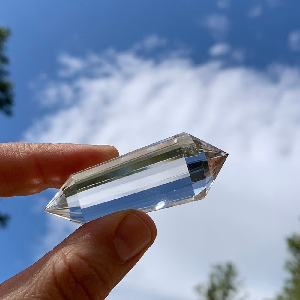 A faceted clear quartz Vogel wand held between finger and thumb against a bright blue sky, light running along its long faces