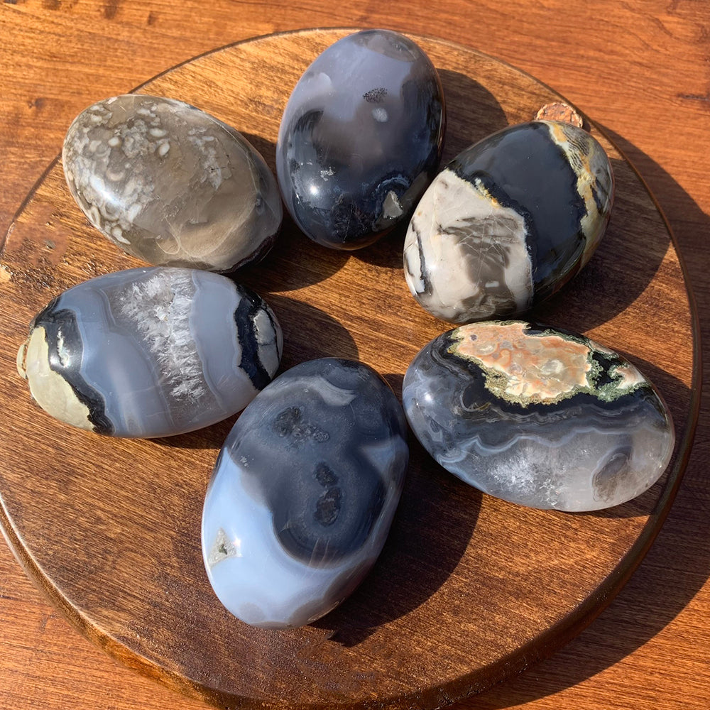 Six polished volcanic agate palmstones in daylight on a wooden board, banded in grey, blue-white and black with earthy rust patches