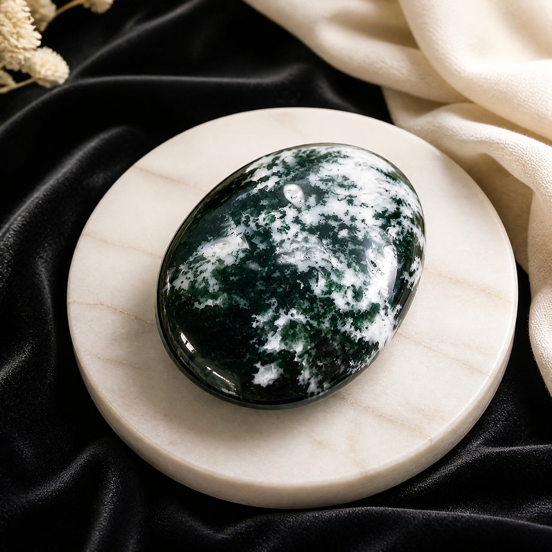 A polished oval green moss agate palmstone with deep green and white moss-like patterns, resting on a pale marble dish